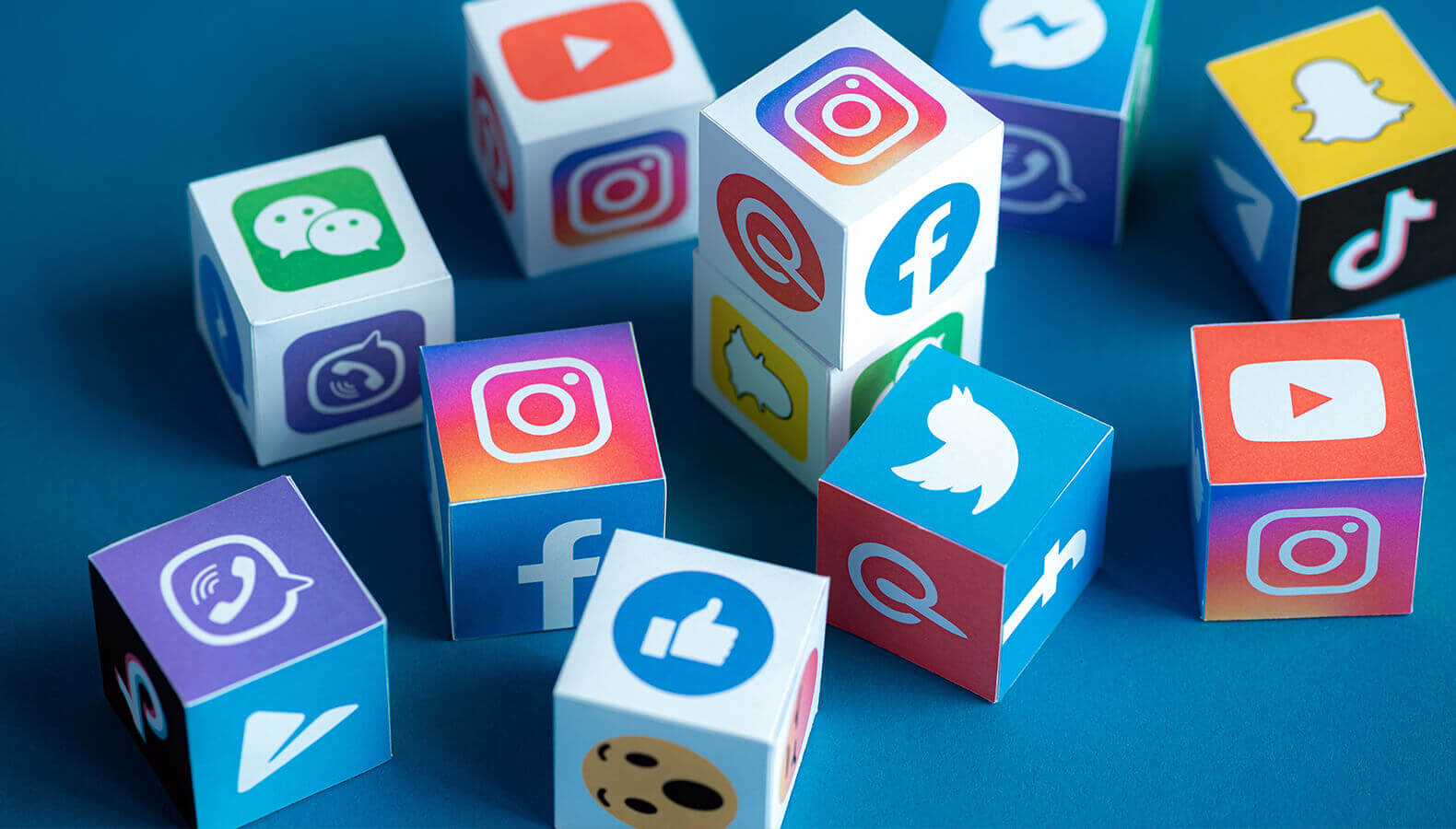 A collection of colorful cubes featuring various social media icons, including Instagram, Facebook, Twitter, YouTube, WhatsApp, WeChat, and Snapchat, scattered on a blue background. Each cube has a different icon on its sides, representing different social media platforms.